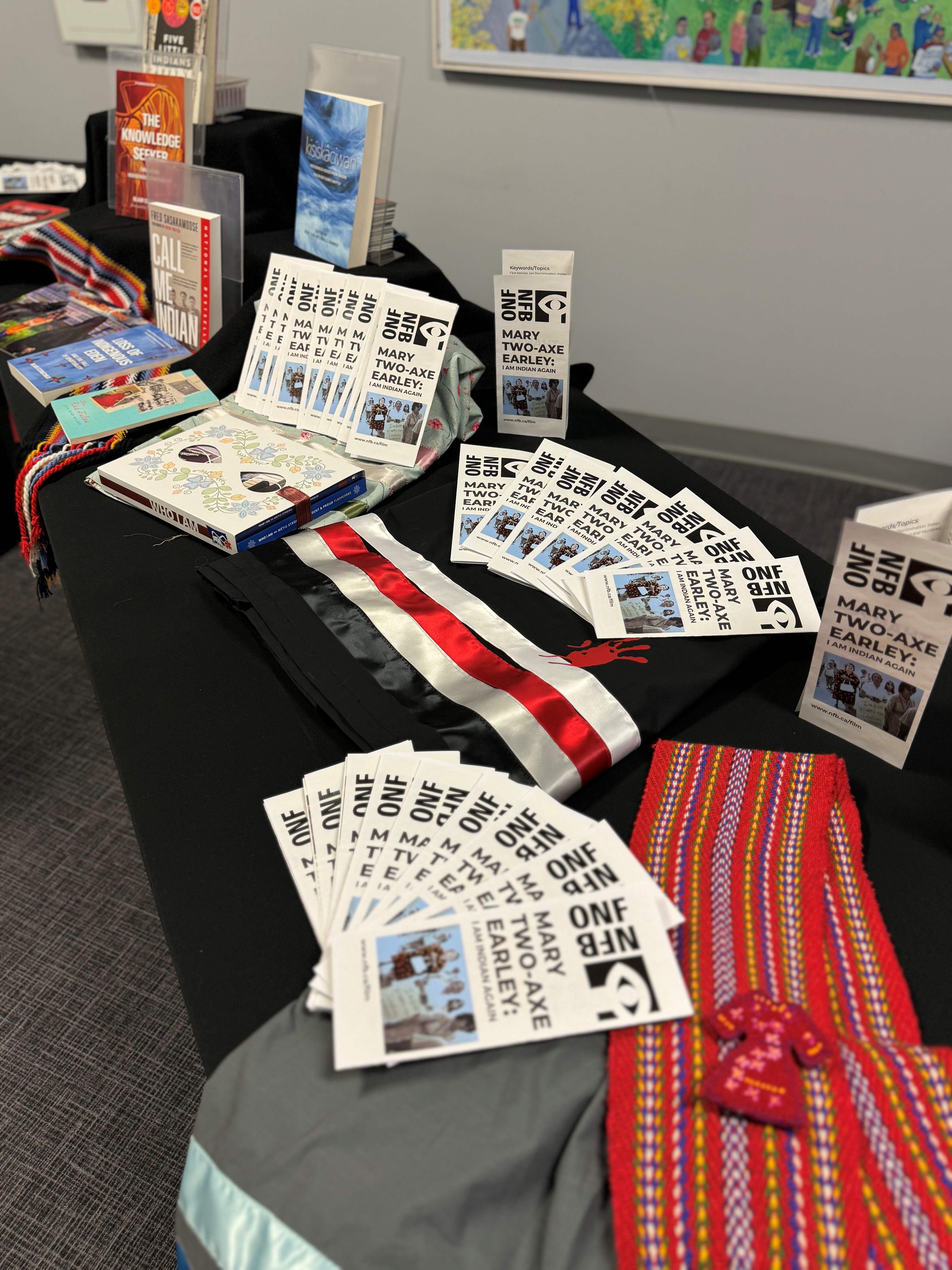 A table display from an Indigenous Committee film night. The table contains National Film Board brochures on the film Mary Two-Axe Earley, as well as an assortment of novels, ribbon skirts and Métis sashes.